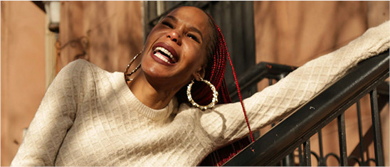 Smiling person wearing hoop earring standing on NYC brownstone stoop.