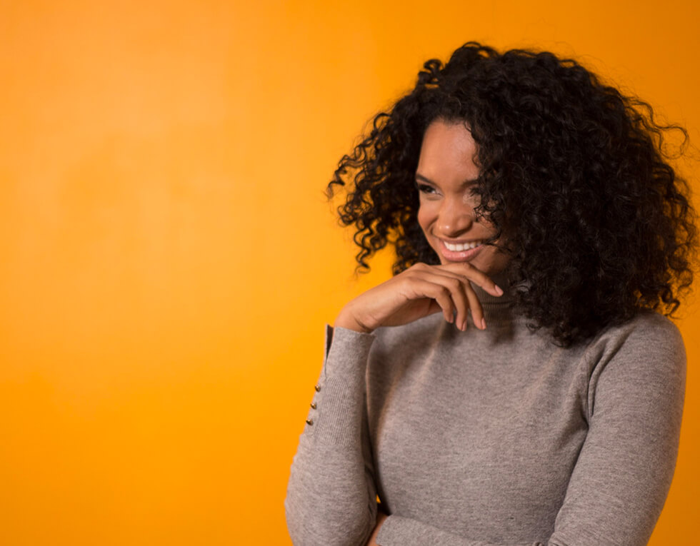 Woman with curly hair and hand on her chin against orange background.