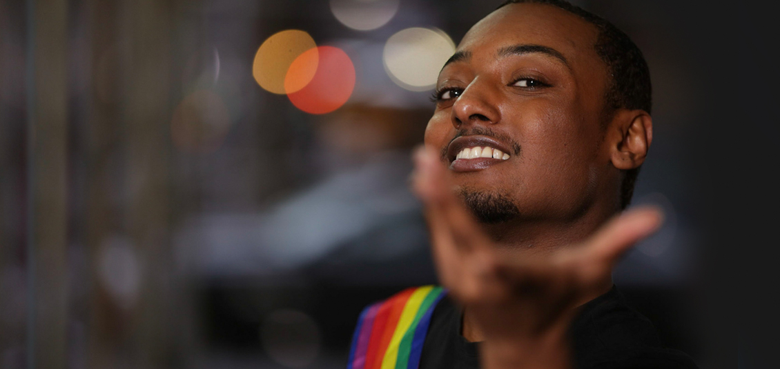 Person wearing a rainbow sash with hand reaching outward to viewers.